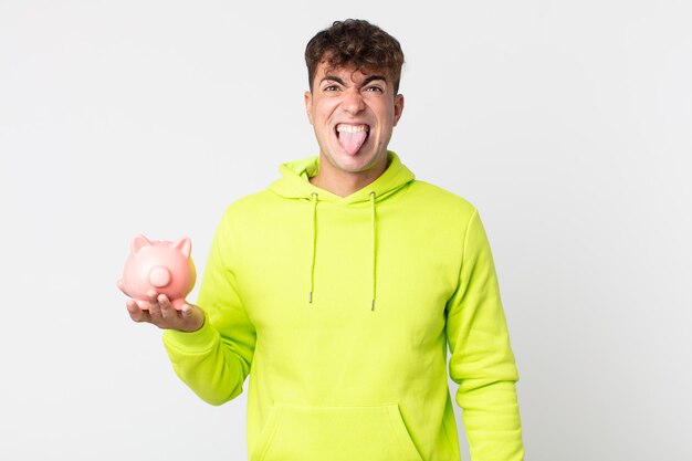 Young handsome man feeling disgusted and irritated and tongue out and holding a piggy bank