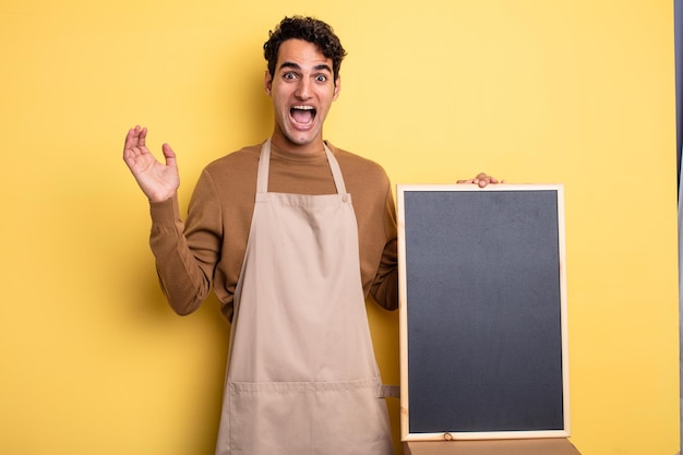 Young handsome man feeling happy and astonished at something unbelievable. chef and blackboard concept