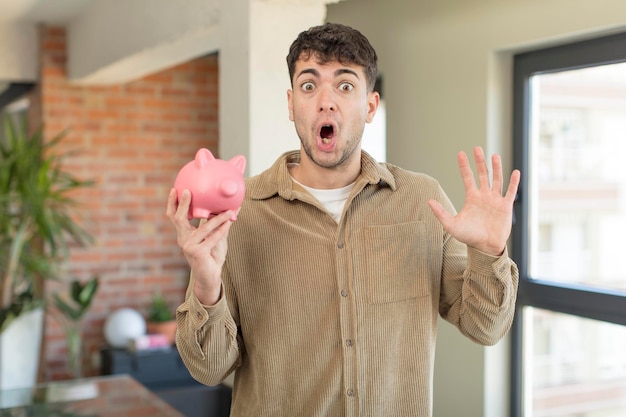 young handsome man feeling happy and astonished at something unbelievable. piggy bank concept