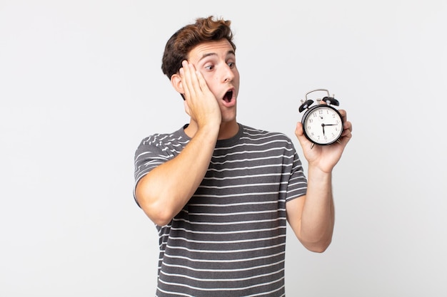 Young handsome man feeling happy, excited and surprised and holding an alarm clock
