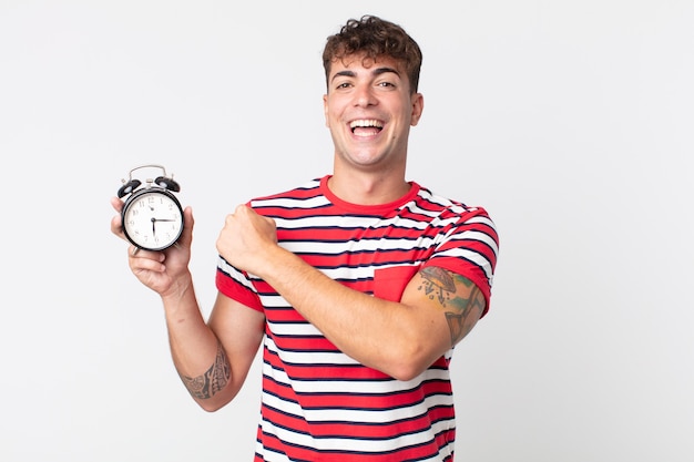 Young handsome man feeling happy and facing a challenge or celebrating and holding an alarm clock