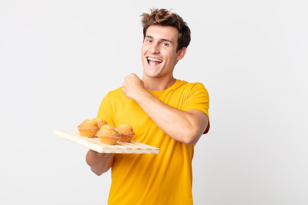 Young handsome man feeling happy and facing a challenge or celebrating holding a muffins tray