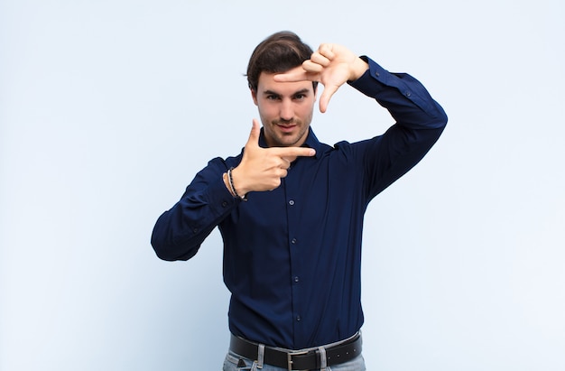 Young handsome man feeling happy, friendly and positive, smiling and making a portrait or photo frame with hands against blue wall