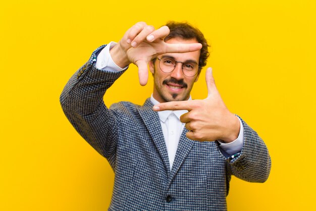 Giovane uomo bello sentirsi felice, amichevole e positivo, sorridendo e facendo un ritratto o una cornice con le mani contro la parete arancione