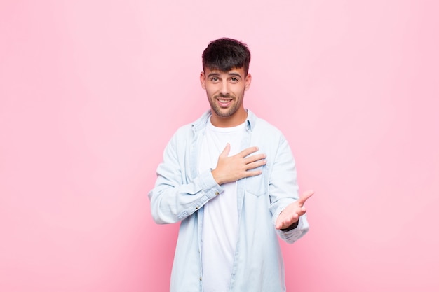 Young handsome man feeling happy and in love, smiling with one hand next to heart and the other stretched up front against pink wall