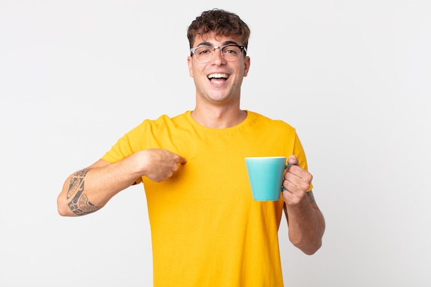 幸せを感じ、興奮してコーヒーカップを持って自己を指している若いハンサムな男