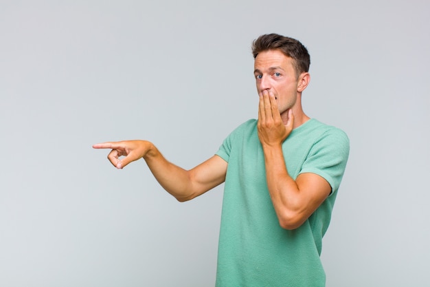 Young handsome man feeling happy, shocked and surprised, covering mouth with hand and pointing to lateral copy space