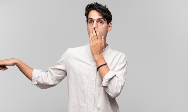 Young handsome man feeling happy, shocked and surprised, covering mouth with hand and pointing to lateral copy space