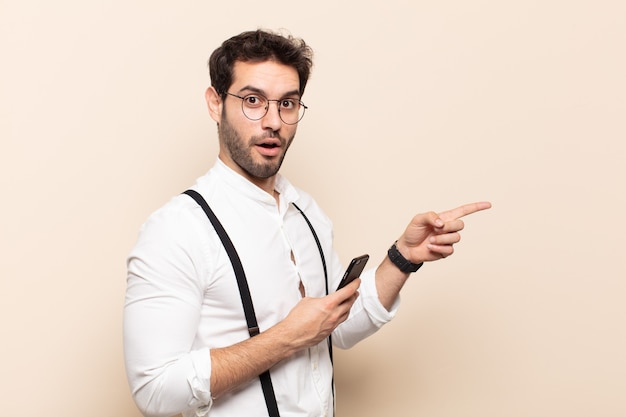 Young handsome man feeling happy, shocked and surprised, covering mouth with hand and pointing to lateral copy space