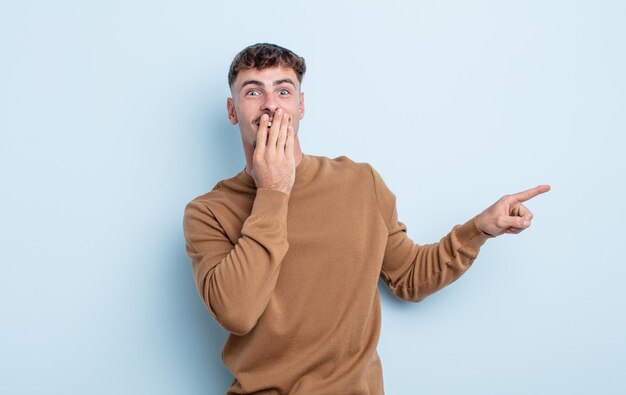 Young handsome man feeling happy, shocked and surprised, covering mouth with hand and pointing to lateral copy space