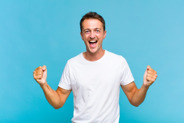 Young handsome man feeling happy, surprised and proud, shouting and celebrating success with a big smile