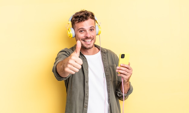 Giovane uomo bello sentirsi orgoglioso, sorridendo positivamente con il pollice in alto le cuffie e il concetto di smartphone