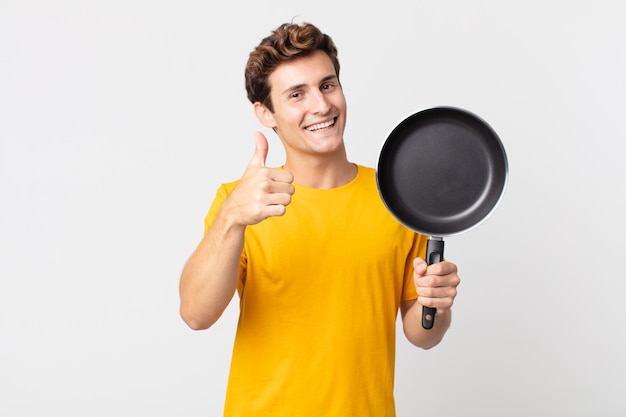 Young handsome man feeling proud,smiling positively with thumbs up and holding a cook pan