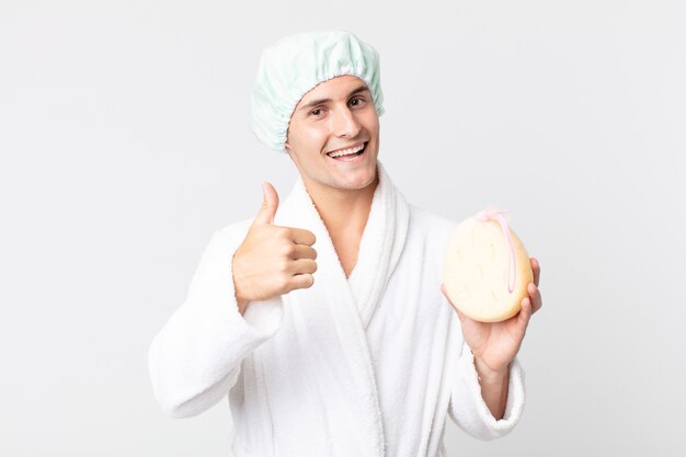 Young handsome man feeling proud,smiling positively with thumbs up with bathrobe, shower cap and a sponge