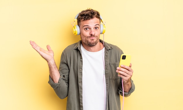 Young handsome man feeling puzzled and confused and doubting headphones and smartphone concept