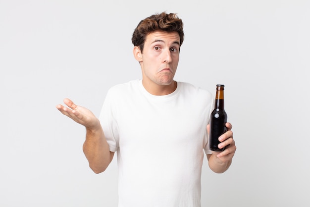 Young handsome man feeling puzzled and confused and doubting and holding a beer bottle