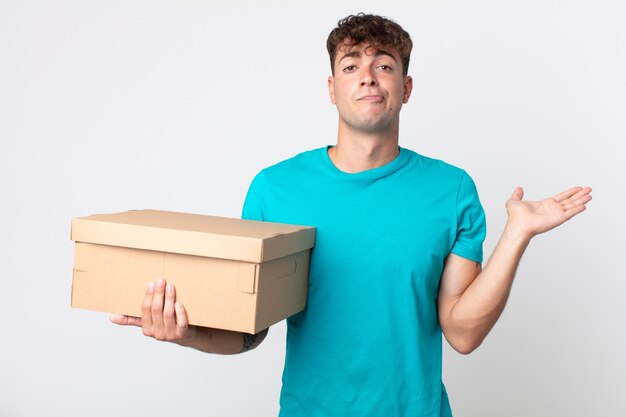 Young handsome man feeling puzzled and confused and doubting and holding a cardboard box