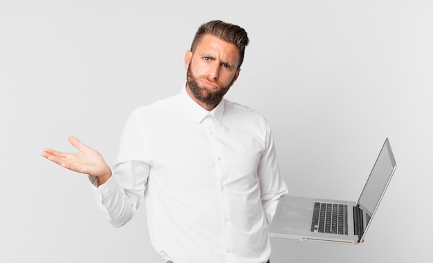 Young handsome man feeling puzzled and confused and doubting and holding a laptop