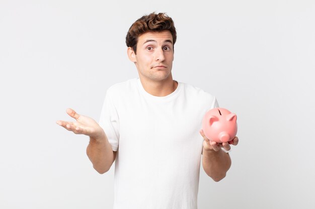 Young handsome man feeling puzzled and confused and doubting and holding a piggy bank