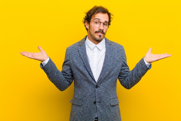 Young handsome man feeling puzzled and confused, doubting, weighting or choosing different options with funny expression against orange wall