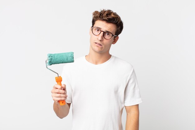 Young handsome man feeling puzzled and confused and holding a roller paint