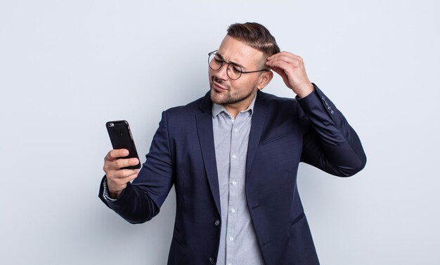 Young handsome man feeling puzzled and confused, scratching head business and smartphone concept