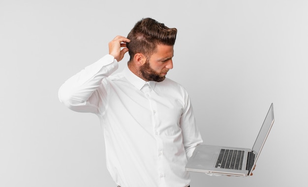 Young handsome man feeling puzzled and confused, scratching head and holding a laptop