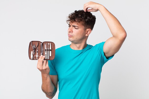 Young handsome man feeling puzzled and confused, scratching\
head and holding a nails tools case