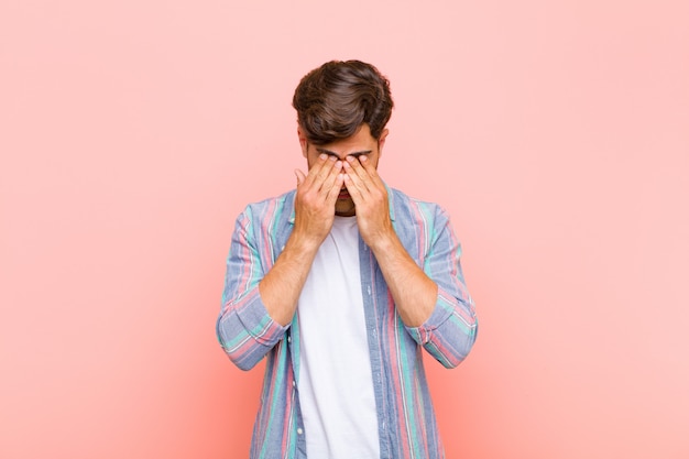 Young handsome man feeling sad, frustrated, nervous and depressed, covering face with both hands, crying against pink wall