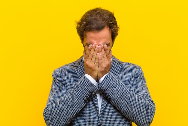 Young handsome man feeling sad, frustrated, nervous and depressed, covering face with both hands, crying  orange wall