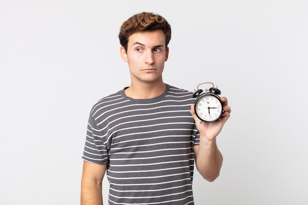 Young handsome man feeling sad upset or angry and looking to the side and holding an alarm clock