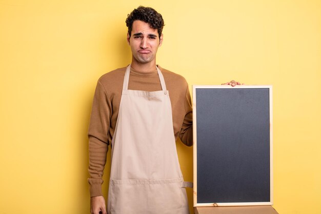 Young handsome man feeling sad and whiney with an unhappy look and crying. chef and blackboard concept