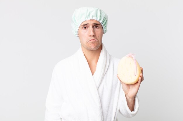 Young handsome man feeling sad and whiney with an unhappy look and crying with bathrobe, shower cap and a sponge