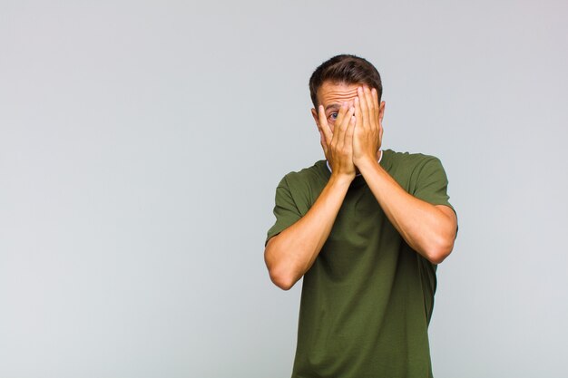 Young handsome man feeling scared or embarrassed, peeking or spying with eyes half-covered with hands