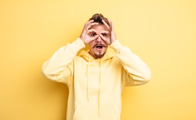 Young handsome man feeling shocked, amazed and surprised, holding glasses with astonished, disbelieving look. strange moustache concept