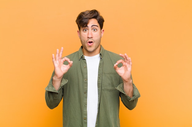 Young handsome man feeling shocked, amazed and surprised, showing approval making okay sign with both hands against orange wall