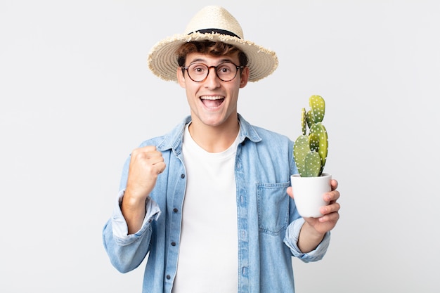 Young handsome man feeling shocked,laughing and celebrating success. farmer holding a decorative cactus