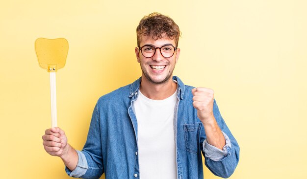 Young handsome man feeling shocked,laughing and celebrating success. flyswatter concept