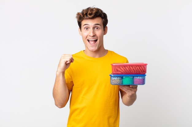 Young handsome man feeling shocked,laughing and celebrating success and holding lunch boxes