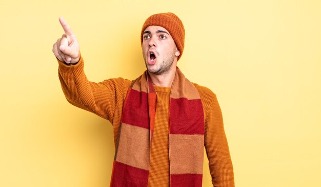 Young handsome man feeling shocked and surprised, pointing and looking upwards in awe with amazed, open-mouthed look