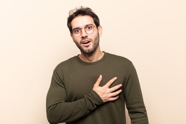 Young handsome man feeling shocked and surprised, smiling, taking hand to heart