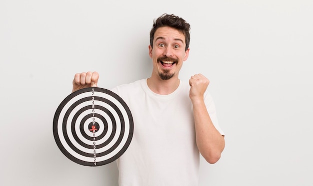 Young handsome man feeling shockedlaughing and celebrating success dart target concept