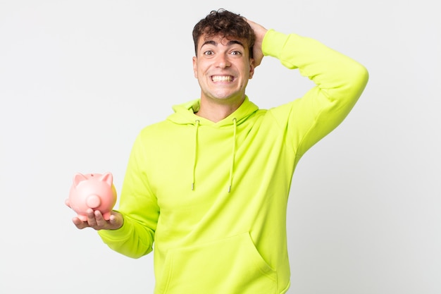 Young handsome man feeling stressed, anxious or scared, with hands on head and holding a piggy bank