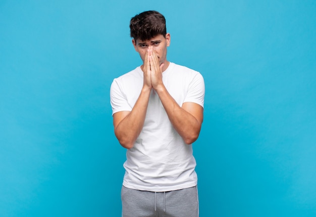 Young handsome man feeling worried, hopeful and religious, praying faithfully with palms pressed, begging forgiveness