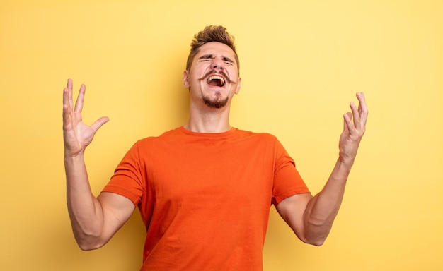 Young handsome man furiously screaming, feeling stressed and annoyed with hands up in the air saying why me. strange moustache concept