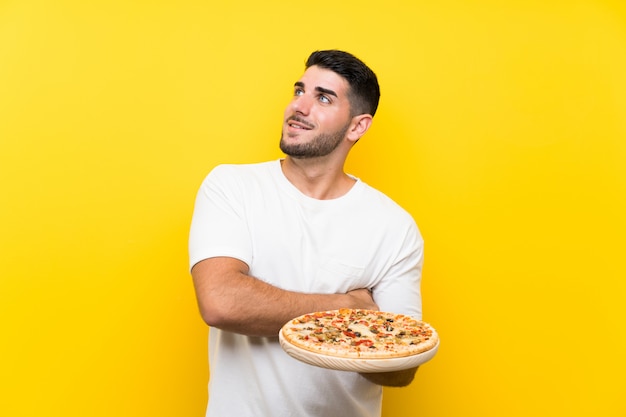 Молодой красивый мужчина держит пиццу на изолированной желтой стене, глядя вверх, в то время как улыбается