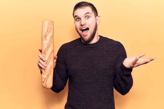 Young handsome man holding baguette celebrating achievement with happy smile and winner expression with raised hand