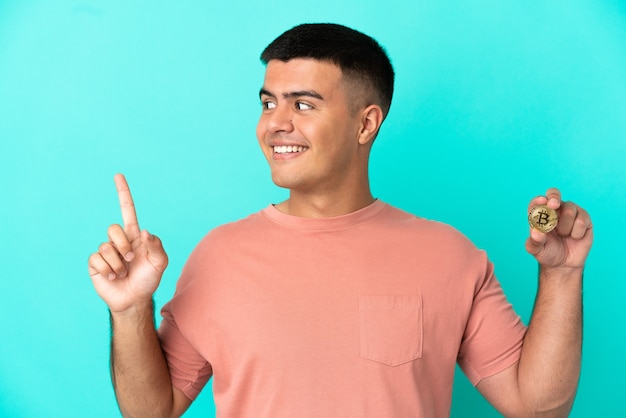 Young handsome man holding a Bitcoin over isolated blue background pointing up a great idea