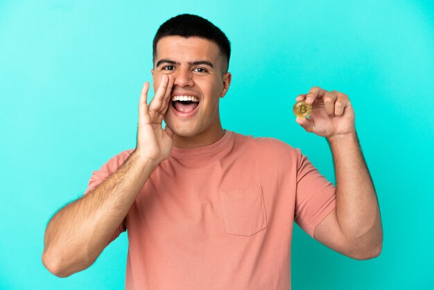 Young handsome man holding a Bitcoin over isolated blue background shouting with mouth wide open