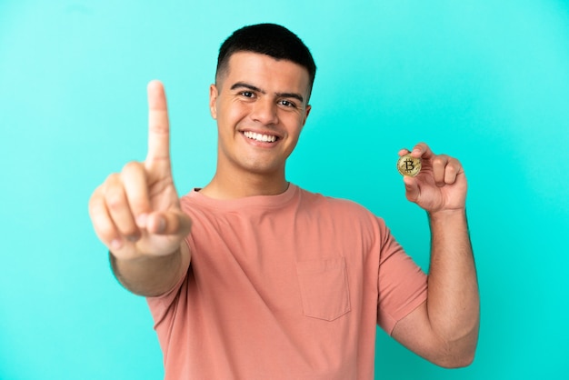 Foto giovane uomo bello che tiene un bitcoin su sfondo blu isolato che mostra e solleva un dito